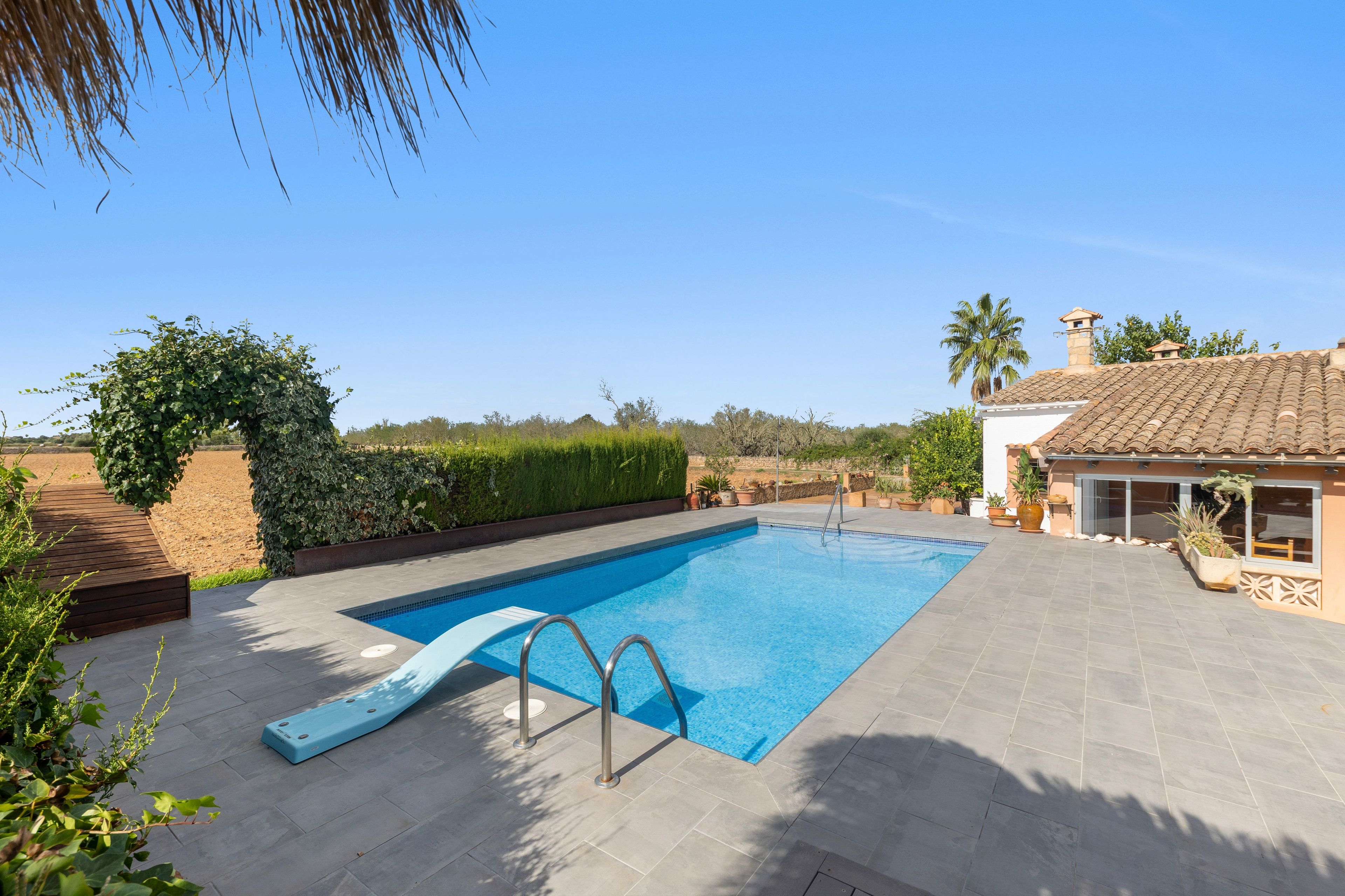 A rectangular swimming pool with a slide, surrounded by a paved deck, with a building and vegetation in the background.