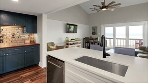 Une cuisine moderne avec un plan de travail blanc, des meubles de couleur bleu foncé et un lave-vaisselle. On y trouve un coin repas avec une table et des chaises, et une vue sur l’océan grâce à de grandes fenêtres.