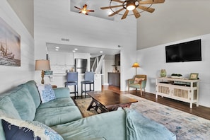 A spacious living room with a sofa, a coffee table, and a TV mounted on the wall. There is a kitchen area in the background with white cabinets and a dining table.