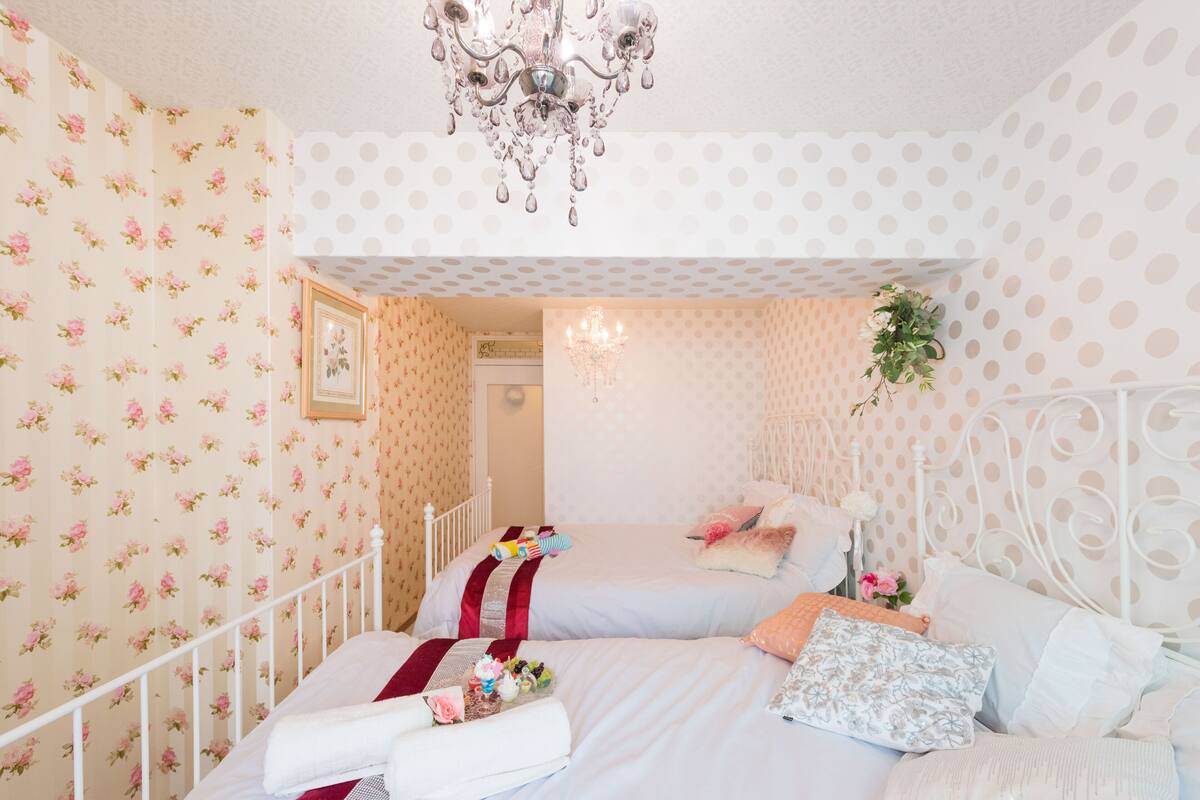 Une chambre avec deux lits, un papier peint à motifs floraux, un lustre et un cadre de lit en métal blanc.