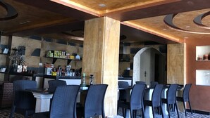 A restaurant with a modern interior, featuring a bar area with shelves stocked with bottles and glasses, and several black chairs arranged around tables.