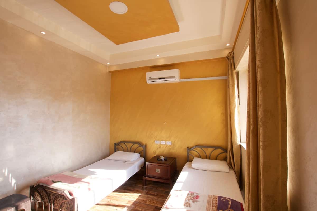 A hotel room with two single beds, an air conditioning unit, a small wooden nightstand, and a window with curtains.