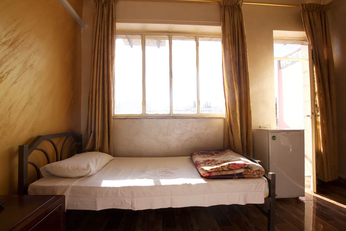 A single bed with a headboard, a small bedside table, a nightstand, a window with curtains, and a refrigerator.