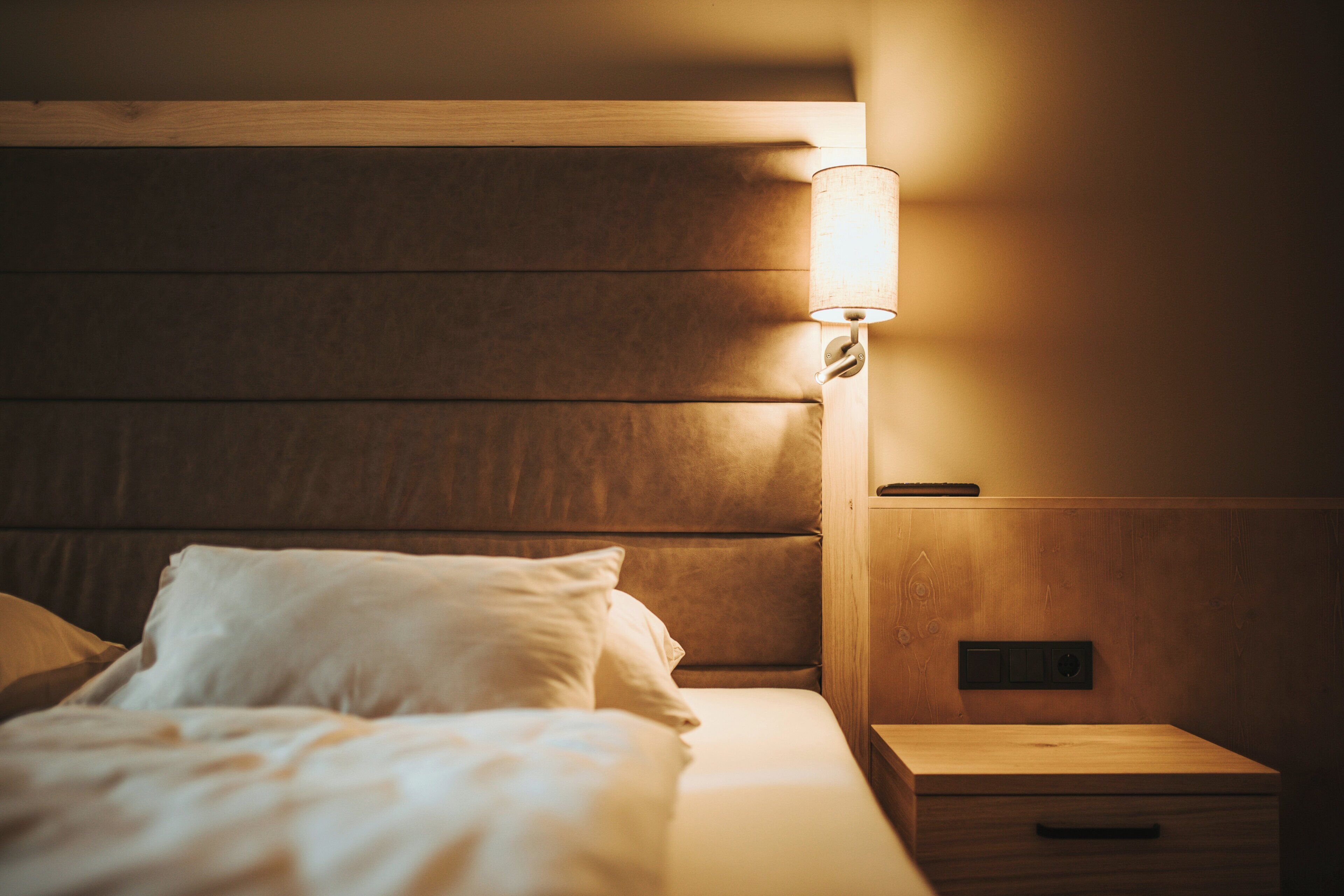 A neatly made bed with a headboard, a bedside lamp, and a wooden nightstand with a drawer.