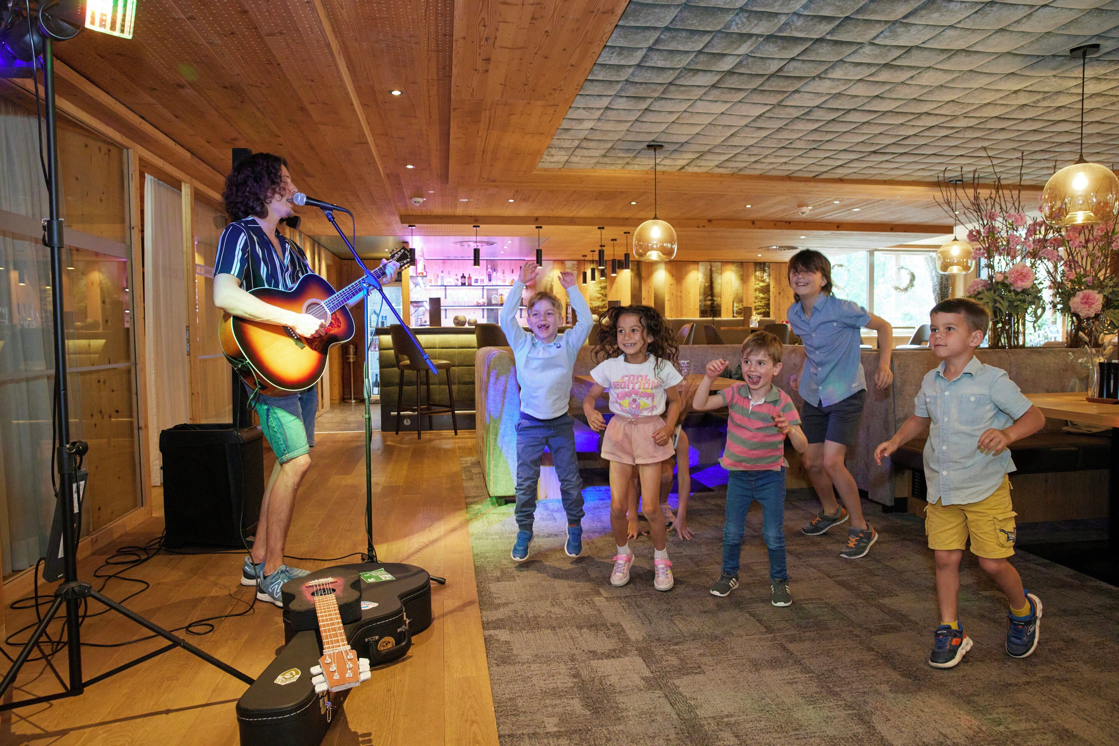 A musician performing with a guitar and a microphone, surrounded by children and adults in a room with wooden floors and a bar area in the background.