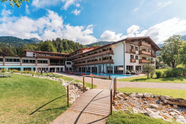 Un hotel de varios pisos con piscina, rodeado de un jardín ajardinado y montañas.