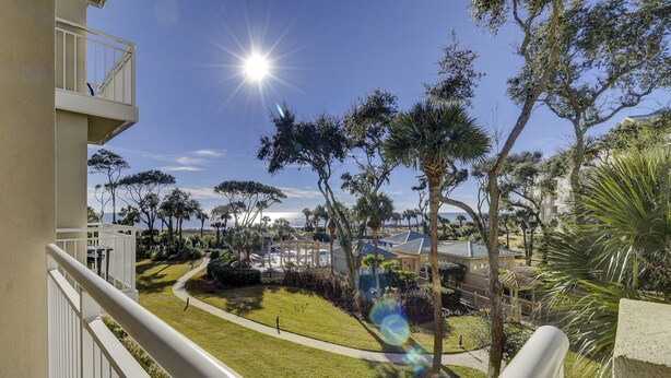 A balcony with a view of a well-maintained garden, a pathway, and a building with a blue roof. - 6204 Hampton Place - Oceanfront Views, Pool, Spa & So Much More! (Hilton Head Island)