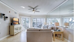 A spacious living room with a beige sofa, a ceiling fan, and large windows offering a view of the outside. - 6204 Hampton Place - Oceanfront Views, Pool, Spa & So Much More! (Hilton Head Island)