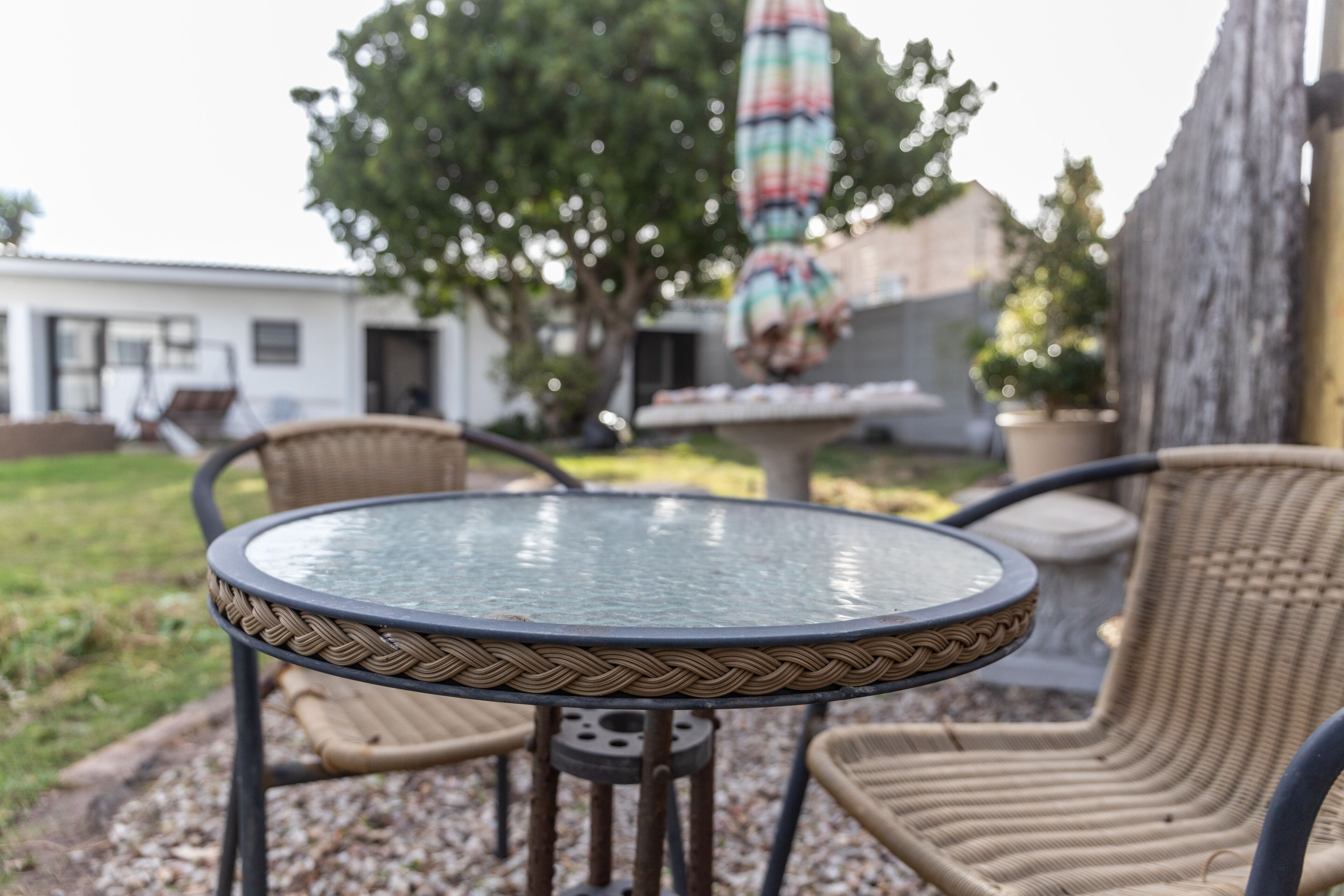 A round glass table with woven metal legs, surrounded by wicker chairs, set outdoors with a tree and a colorful umbrella in the background.