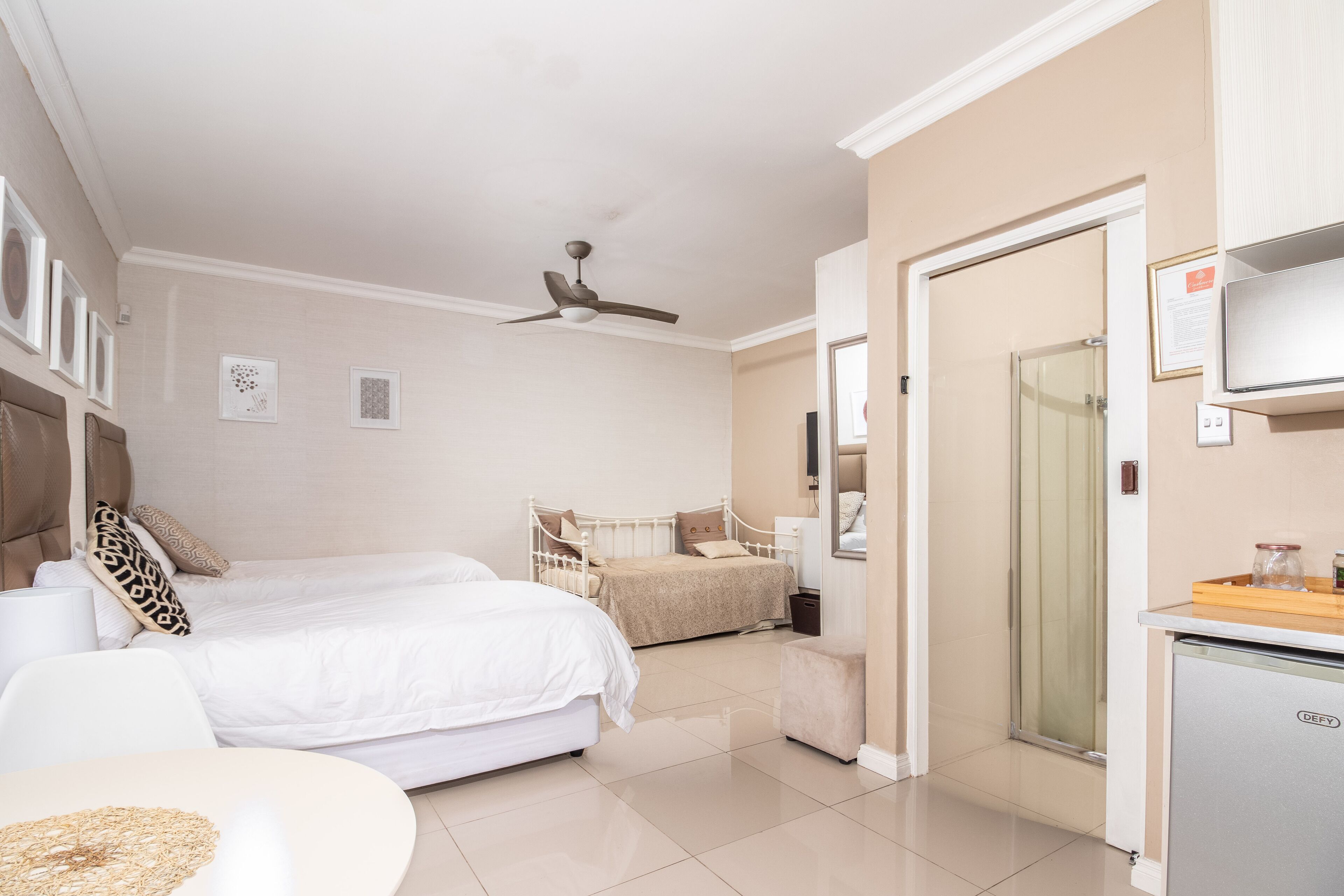 A hotel room with two beds, a ceiling fan, a minibar, and a bathroom with a glass shower.