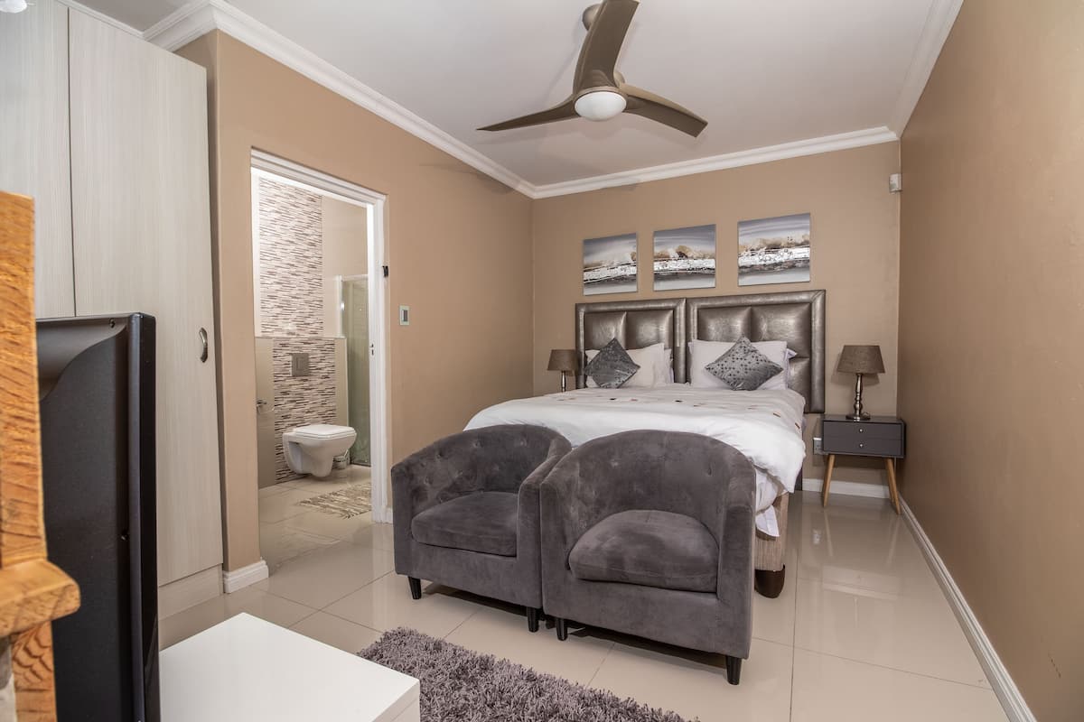 A bedroom with a bed, two armchairs, a nightstand, a ceiling fan, and artwork on the wall.