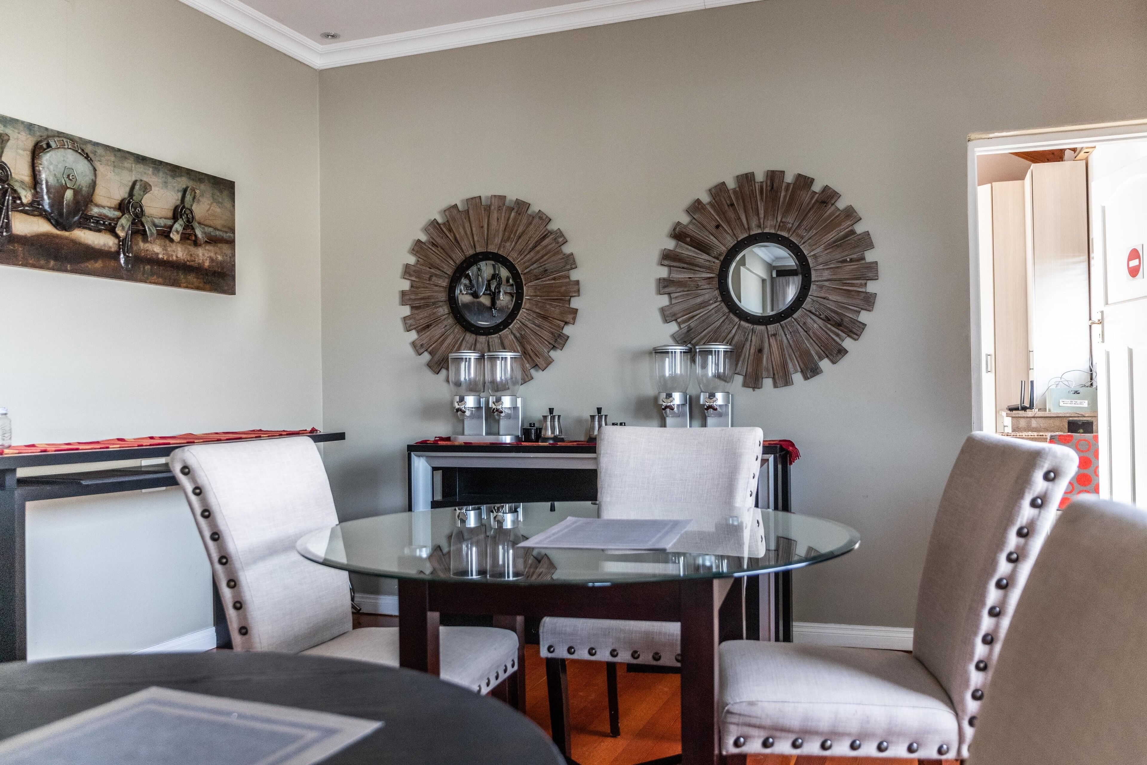 A dining area with a glass table, four upholstered chairs, two wall-mounted mirrors, and a painting above the table.