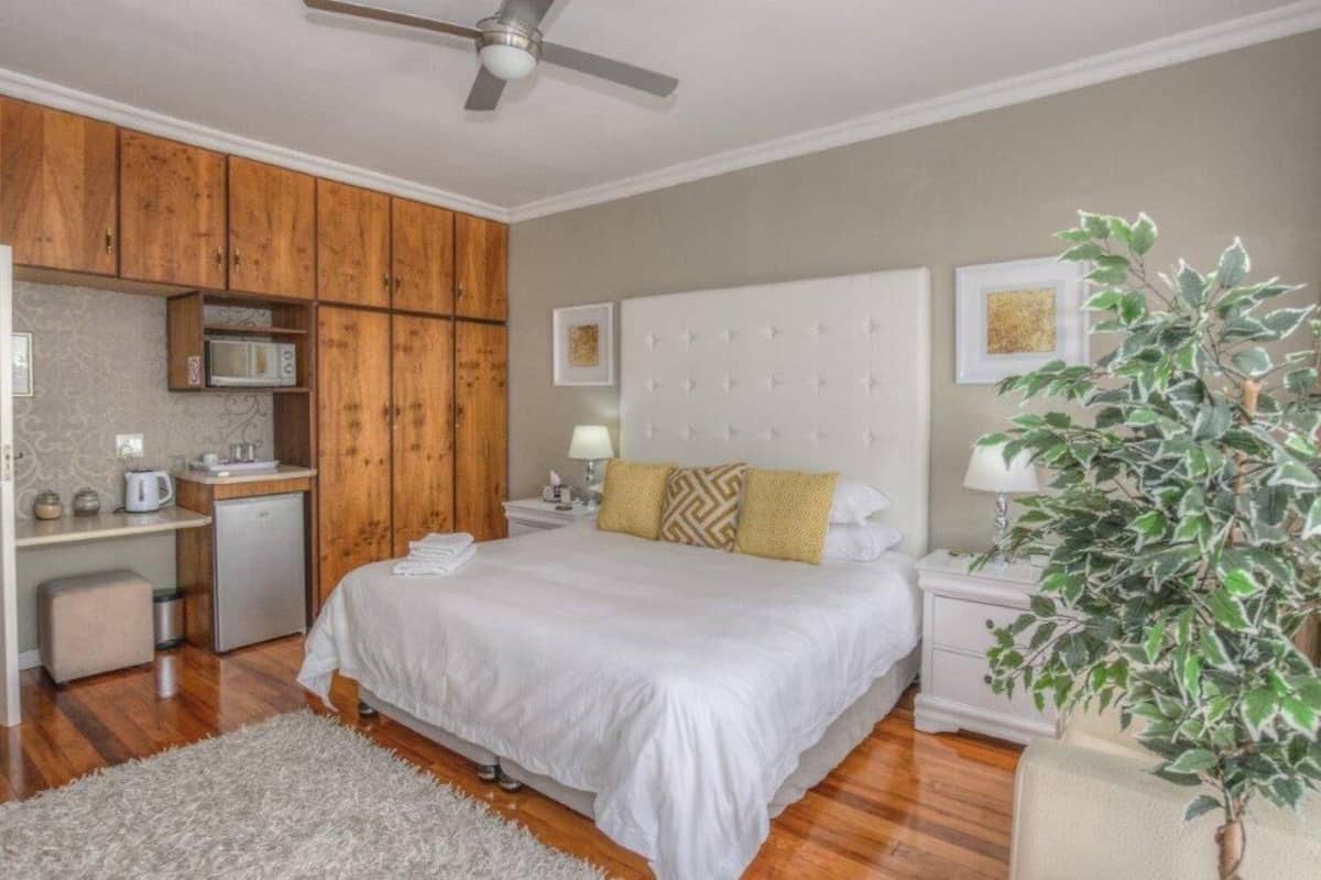 A bedroom with a bed, wooden cabinets, a microwave, a small fridge, a nightstand, a lamp, and a potted plant.