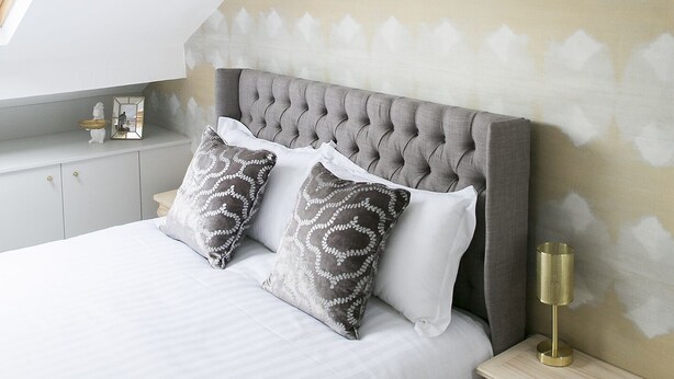 Un dormitorio con cabecera capitonada, cama con ropa blanca, dos almohadas estampadas, mesita de noche con lámpara y una pequeña estantería con adornos.