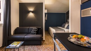 Een knus woongedeelte met een bank, een klein tafeltje met een tijdschrift, een kitchenette en een eettafel met fruit.