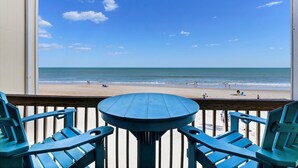 Une vue depuis un balcon, avec une table et des chaises, donnant sur une plage où se trouvent des gens et l’océan.