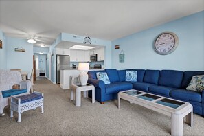 A living room with a blue sofa, a white coffee table, and a wicker chair. There is a kitchen in the background with white cabinets and a microwave.