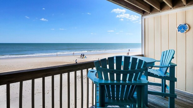Vue sur la plage depuis une terrasse couverte, avec deux chaises Adirondack bleues et une rambarde en bois.