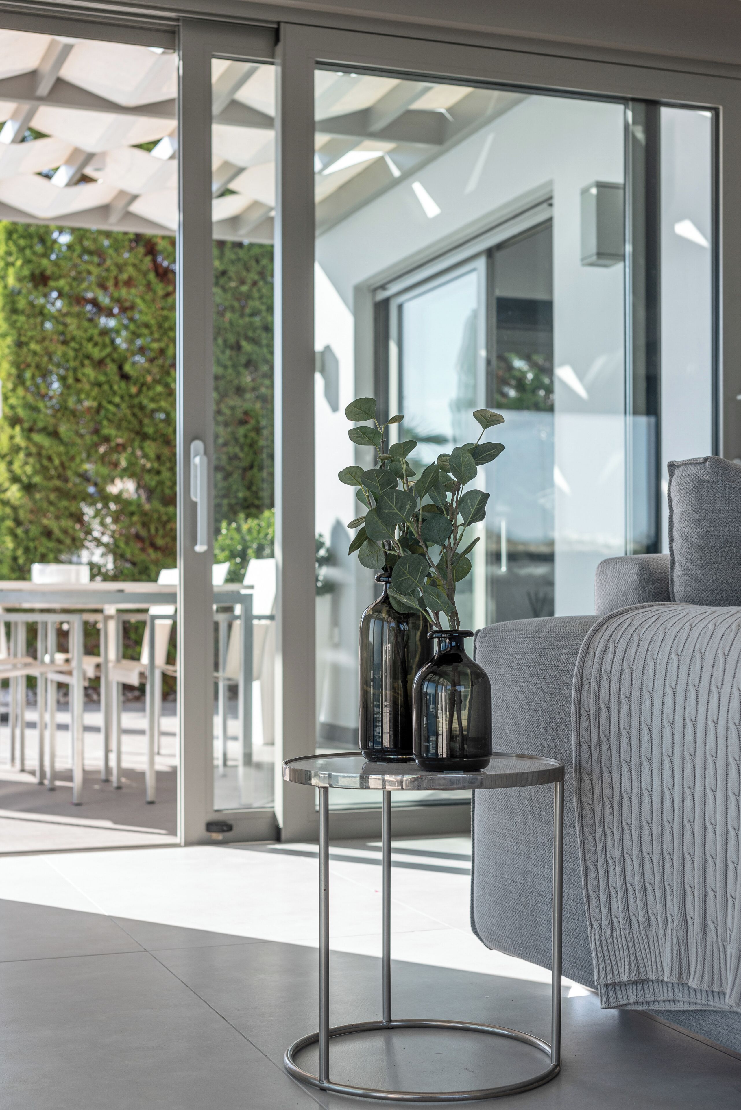 A modern living area with a sofa, a small round table with vases, and a glass door leading to an outdoor seating area.