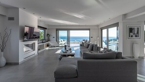 A modern living room with a large gray sofa, a dining area, and a view of the ocean. - Welcome to Villa Soligia! Beach Front Villa Large Private Pool Amazing Sea View (Corinth)