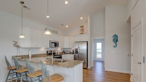 A modern kitchen with white cabinets, granite countertops, and stainless steel appliances. There are four bar stools and a large island with a sink. - Island Oasis, Elevator, Beautiful Decor (Topsail Beach)
