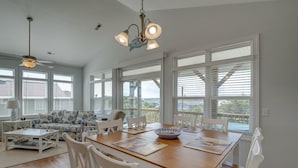 A spacious living room with a wooden dining table, white chairs, a sofa with a blue and white pattern, and large windows offering a view of the outdoors. - Island Oasis, Elevator, Beautiful Decor (Topsail Beach)