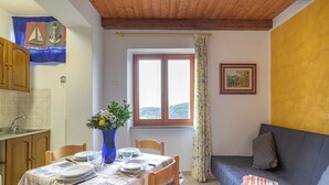 Un comedor con una mesa dispuesta para cuatro personas, un sofá, una ventana con cortinas y una zona de cocina.