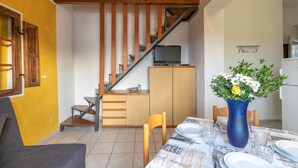 Un comedor con una mesa preparada para dos personas, un jarrón azul con flores, una escalera y un televisor sobre un mueble.