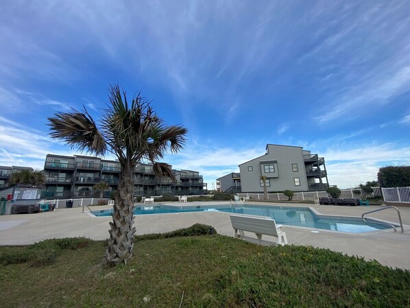 A pool area with a palm tree, a bench, and modern buildings in the background. - Oceanfront Condo with amazing views of The Atlantic (North Topsail Beach)