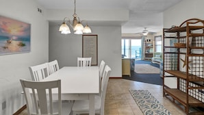 A dining area with a white table and chairs, a chandelier, a framed quote on the wall, and a view to a balcony with a beach scene. - Oceanfront Condo with amazing views of The Atlantic (North Topsail Beach)