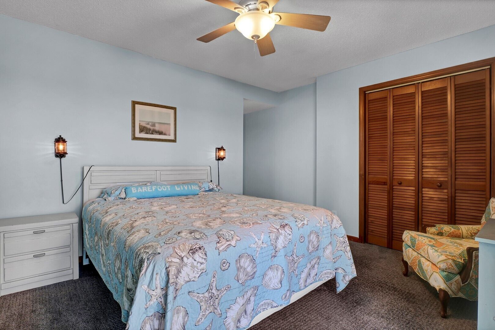A bedroom with a bed, nightstands, a ceiling fan, wall sconces, a wardrobe, and a chair.