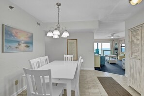 A dining area with a white table and chairs, a chandelier, a painting, and a view of the sea through large windows.