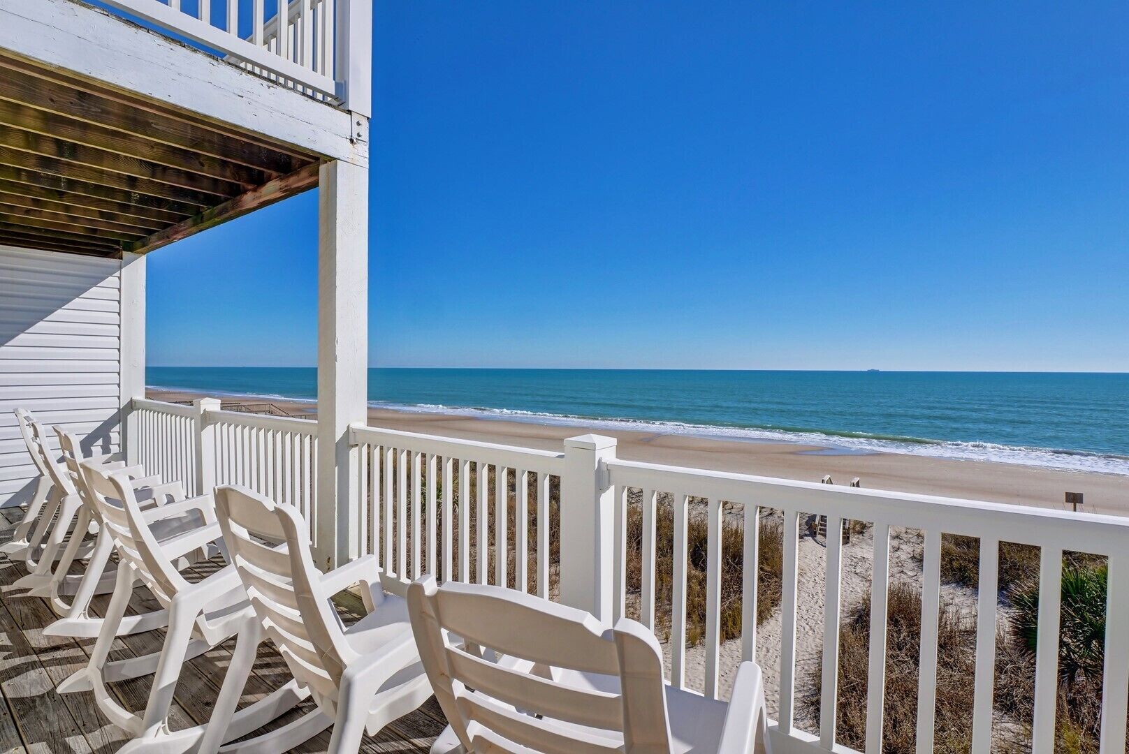 A beachside view from a porch with white railings and beach chairs.