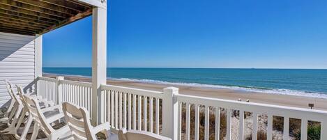 A beachside view from a porch with white railings and beach chairs.
