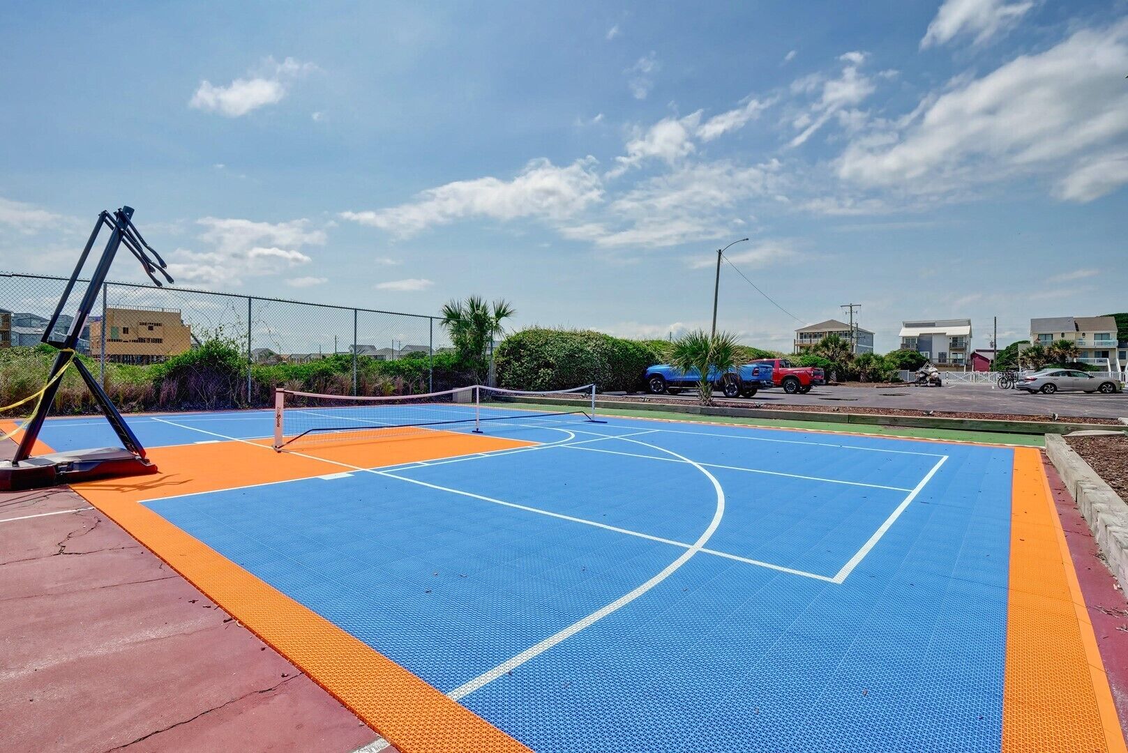An outdoor basketball court with a blue and orange playing surface, surrounded by a fence and palm trees.