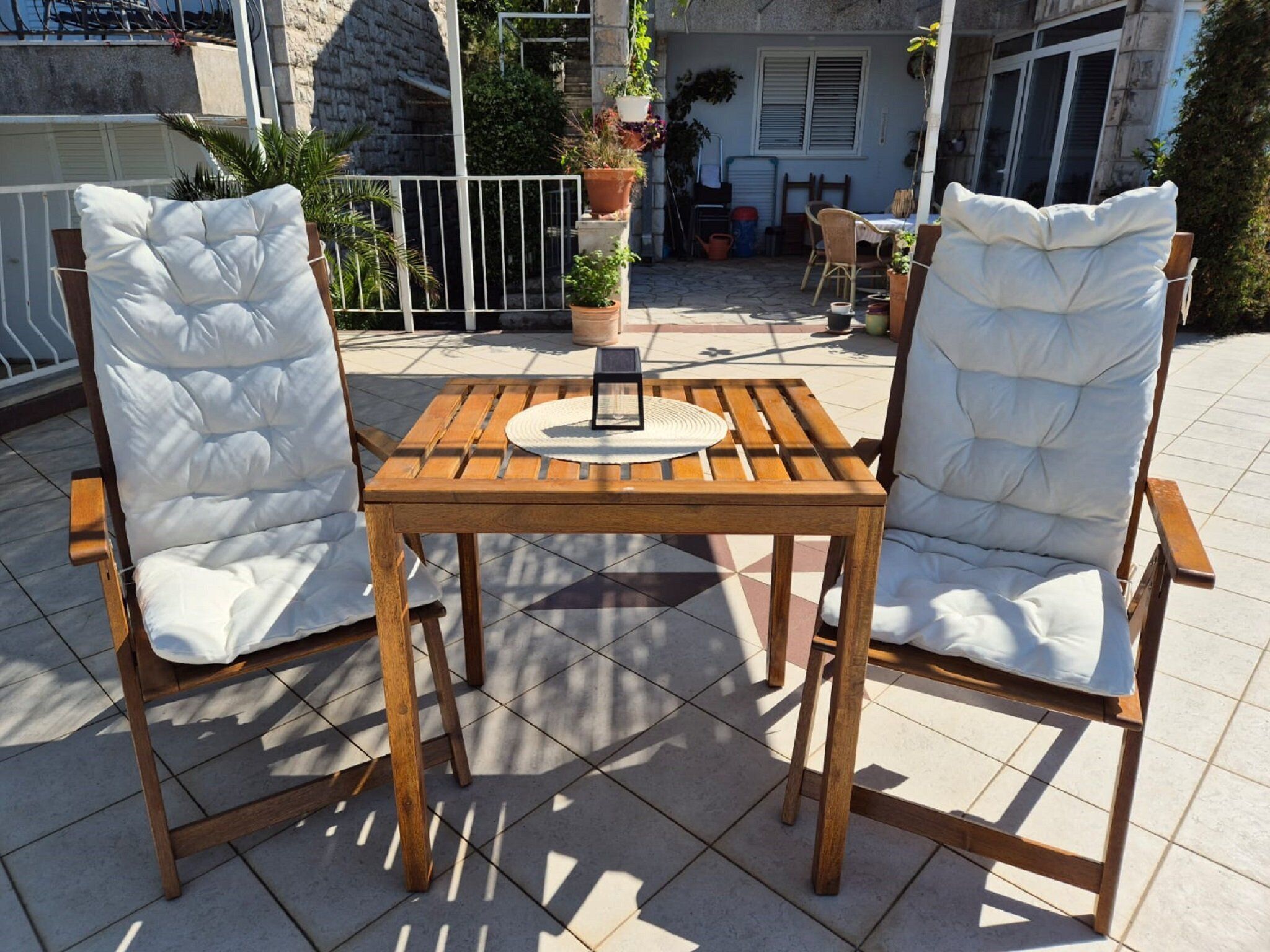 Deux chaises blanches à coussins, dotées d’une structure en bois, et une table en bois aux côtés à lattes, disposées sur une terrasse carrelée agrémentée de plantes en pot, avec un bâtiment en arrière-plan.