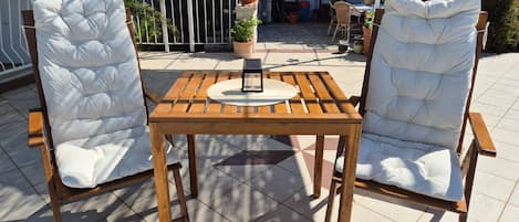 Deux chaises blanches à coussins, dotées d’une structure en bois, et une table en bois aux côtés à lattes, disposées sur une terrasse carrelée agrémentée de plantes en pot, avec un bâtiment en arrière-plan.