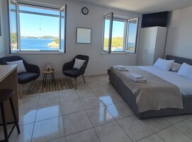 Quarto de hotel com uma cama, duas cadeiras, uma mesinha, um relógio e grandes janelas com vista para o mar.