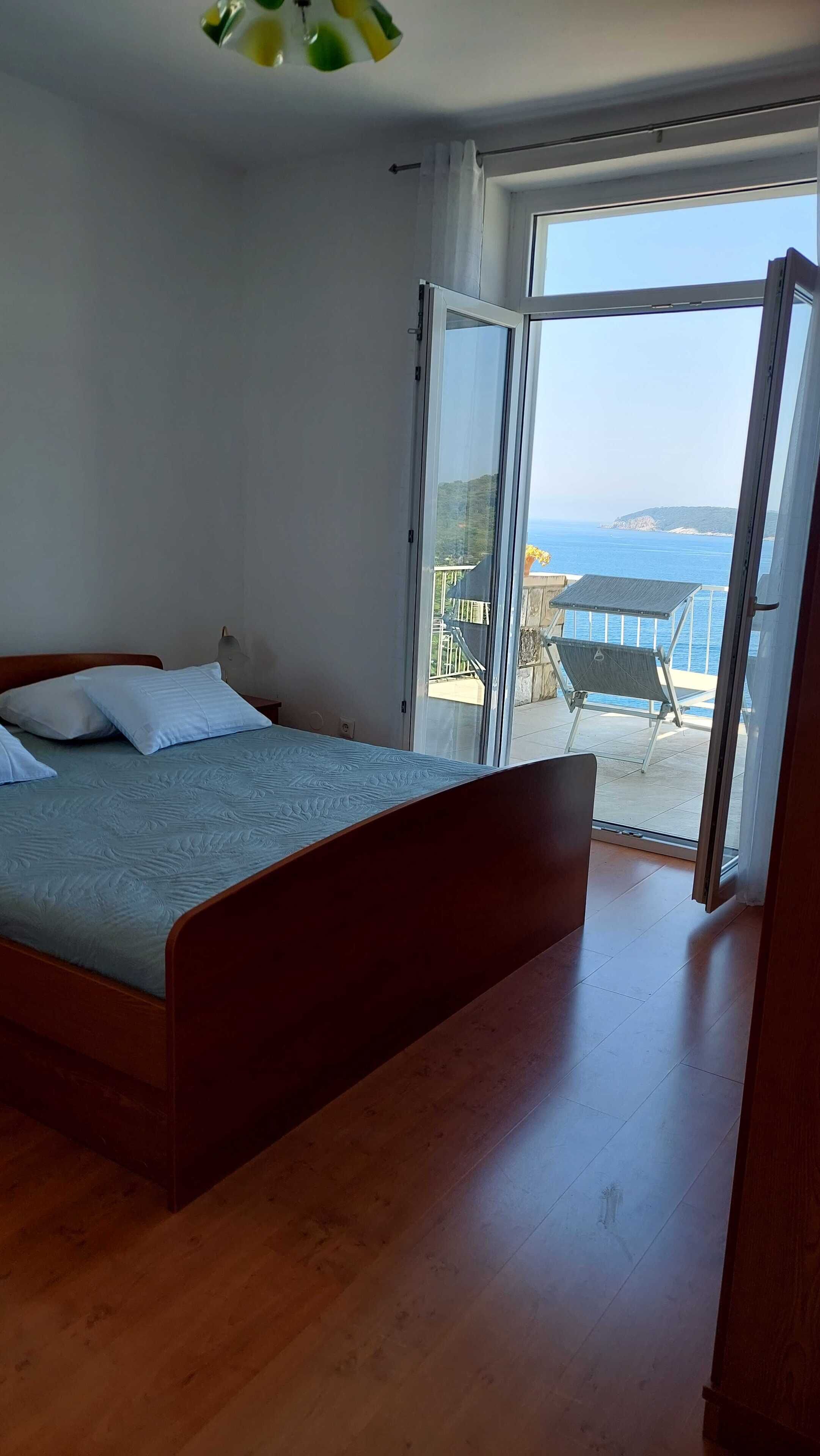 A bedroom with a wooden bed, a large sliding glass door leading to a balcony with a view of the sea.