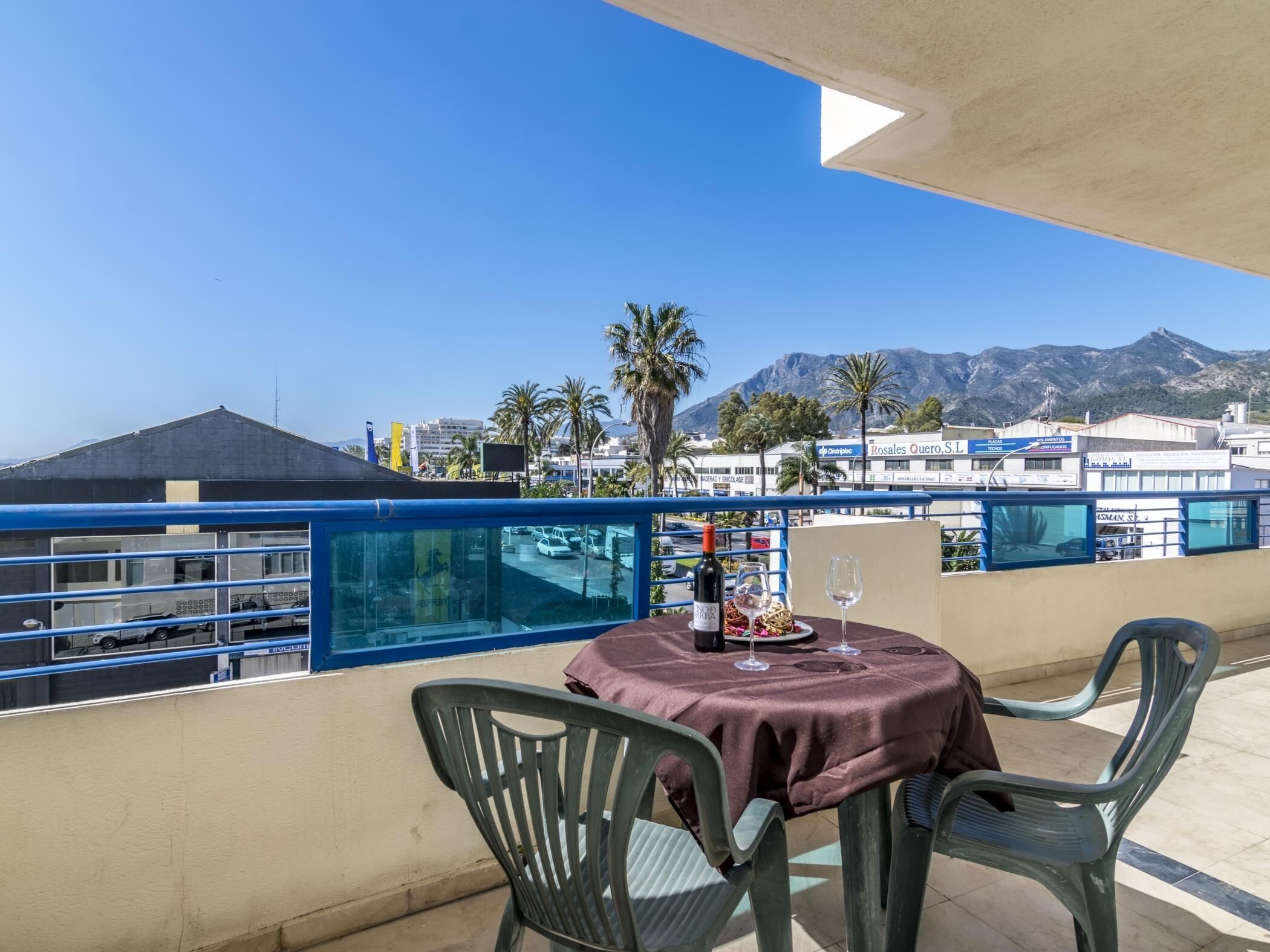 Ein Balkon mit einem Tisch für zwei Personen, mit Blick auf eine Küstenlandschaft mit Gebäuden und Palmen.