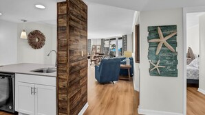 Eine moderne Küche mit weißen Schränken und einer Wand in Holzoptik, ein Essbereich mit einem blauen Sofa und ein Wohnzimmer mit Strandmotiv-Wandbild.