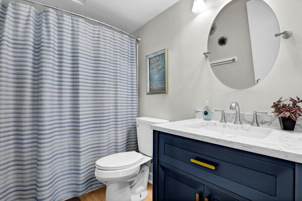 Une salle de bain avec un rideau de douche à rayures, des toilettes, un lavabo avec un plan de travail en marbre, un miroir et un tableau encadré accroché au mur.