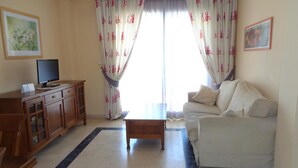 Un salon avec un canapé, une table basse, un téléviseur posé sur un meuble en bois et une fenêtre ornée de rideaux à motifs floraux.