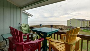 Une terrasse en bois agrémentée de chaises Adirondack aux couleurs vives et d’une petite table, d’où l’on peut admirer une plage et un bâtiment.