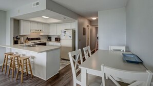 A modern kitchen with white cabinets, a central island with stools, and a dining area with a white table and chairs. - Oceanfront Condo, Steps Away from the beach access (Surf City)