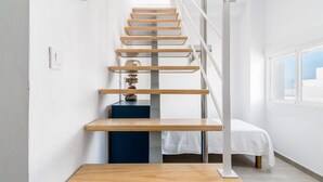 Una escalera de madera con barandales de metal, una cama con sábana blanca y vistas a través de una ventana.