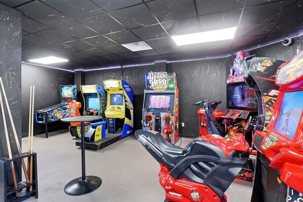 A gaming arcade with multiple racing game machines, a red motorcycle, and a table with chairs.