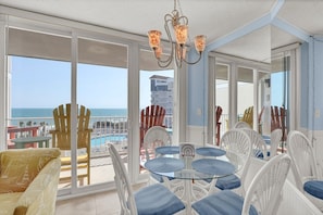 A balcony with a view of the ocean, a pool, and a dining area with chairs and a table.