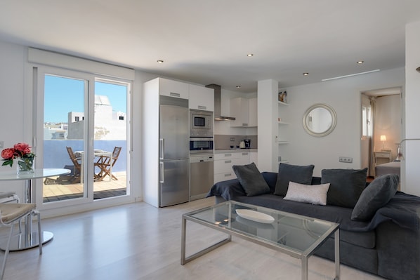 Un appartement moderne doté d’une table basse en verre, d’un canapé gris, d’une cuisine équipée d’appareils électroménagers en acier inoxydable et d’un balcon aménagé avec du mobilier d’extérieur.