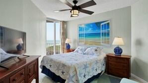 Un dormitorio con una cama, un ventilador de techo, un televisor y un cuadro de una playa.